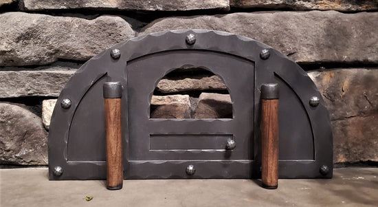 Black iron pizza oven door with wooden handles against a stone wall backdrop