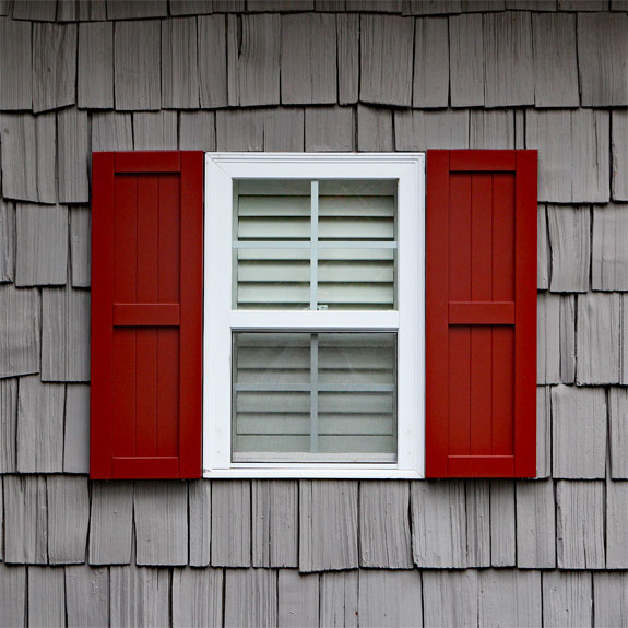Red wooden shutters frame a white window against a rustic gray shingle wall