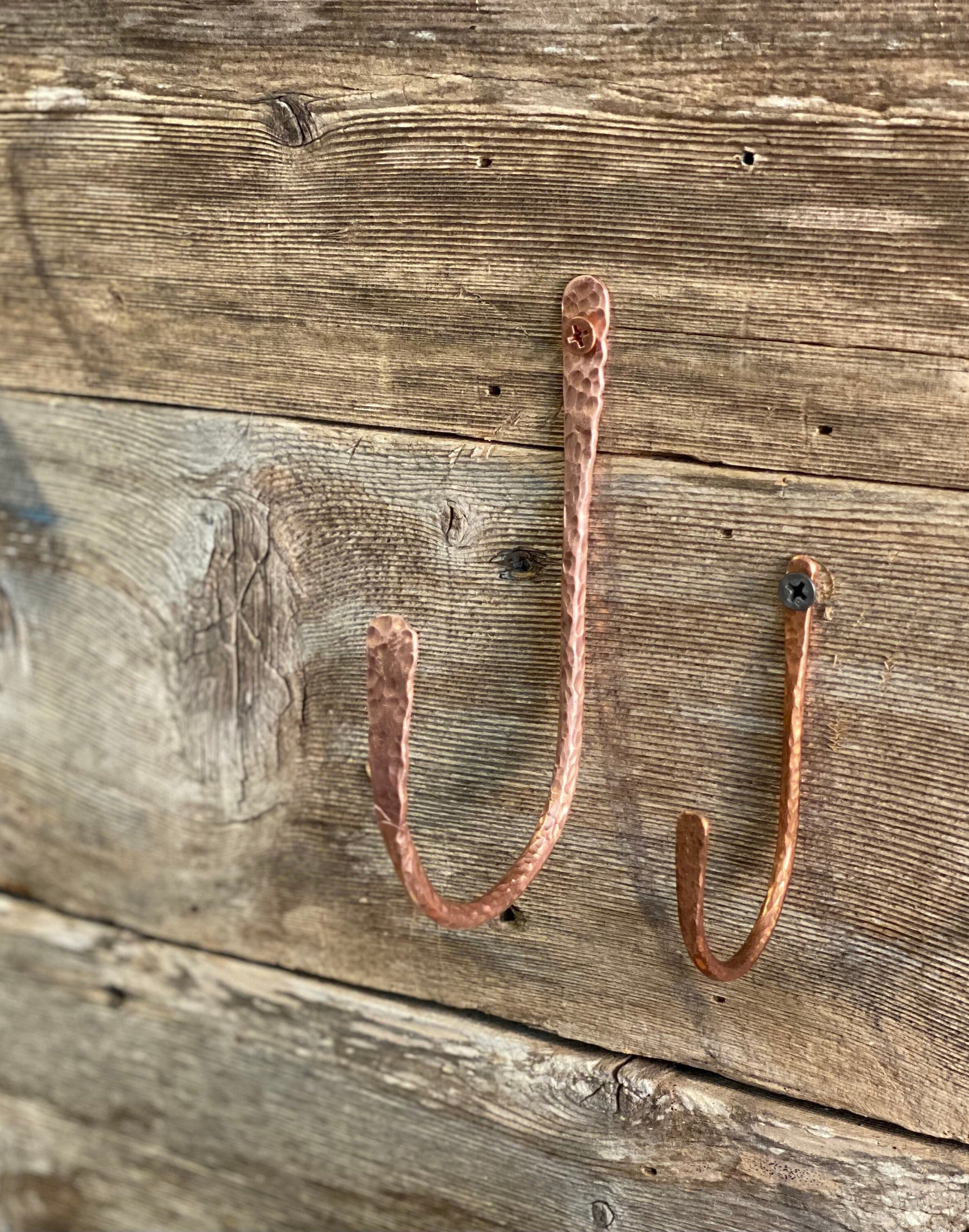 Two hand-hammered J hooks crafted from pure, unfinished copper, mounted on rustic wood. The hooks feature a warm, natural shine and raw metallic luster that will age to a natural patina over time.