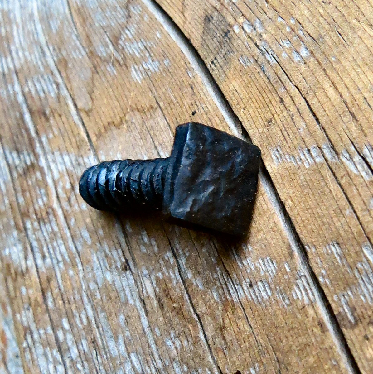A square head lag that has been distressed hammered and painted black lying on a wood background showing off the threads.