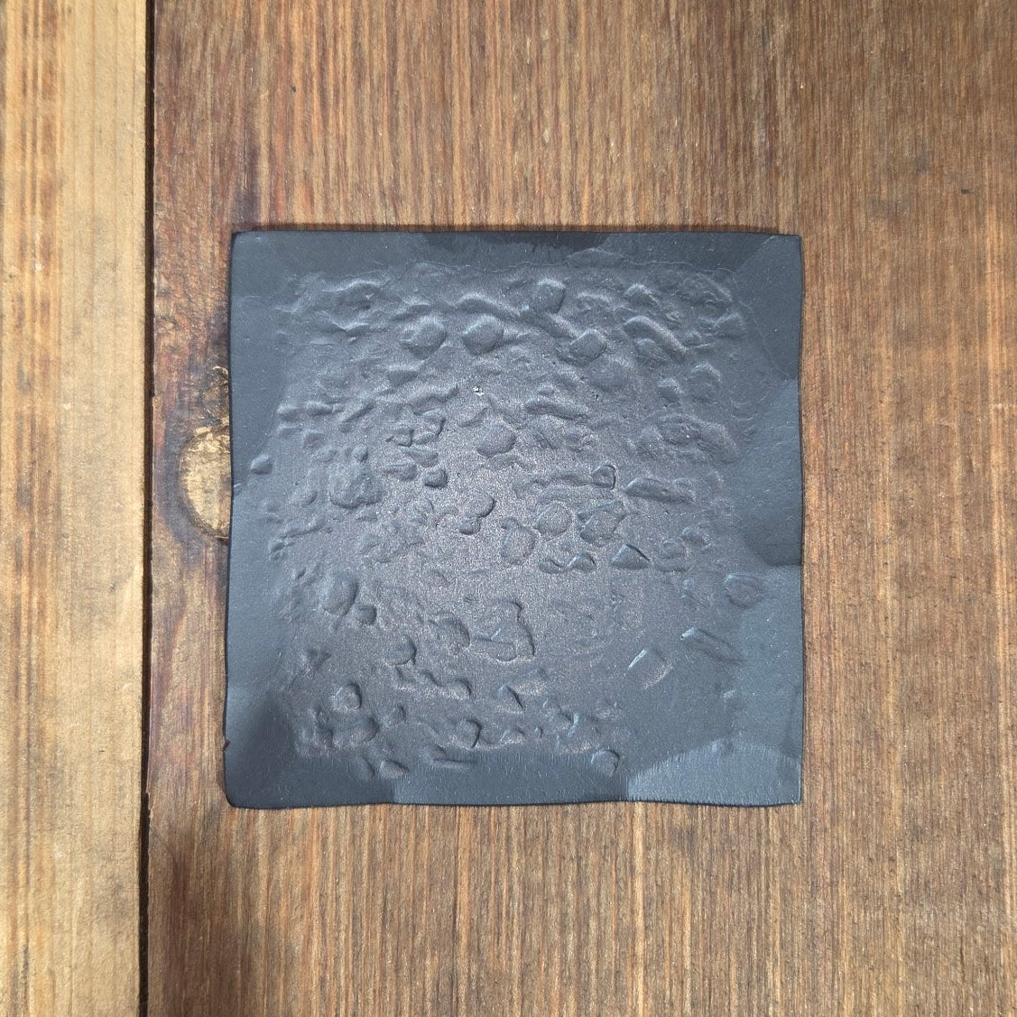 Black square finish and texture sample with an aged hammered design on a wooden surface