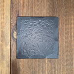 Black square finish and texture sample with an aged hammered design on a wooden surface