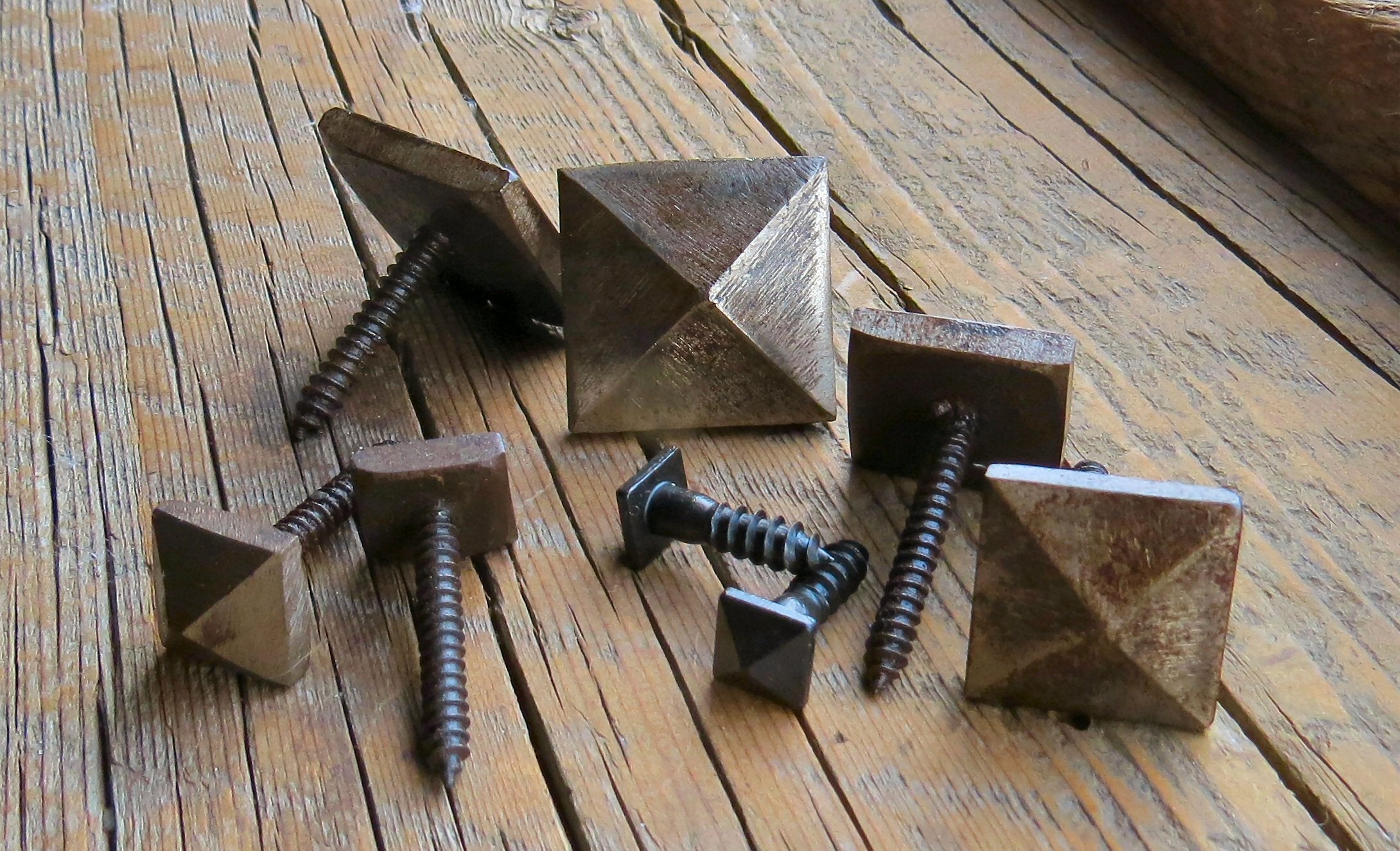 A collection of decorative screws with pyramid heads, displayed on a wooden surface. They have a smooth finish and are shown in various colors including silver, bronze, and black.