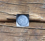 A square head bolt with a smooth texture painted black and finished with aged pewter patina installed on a wood background.