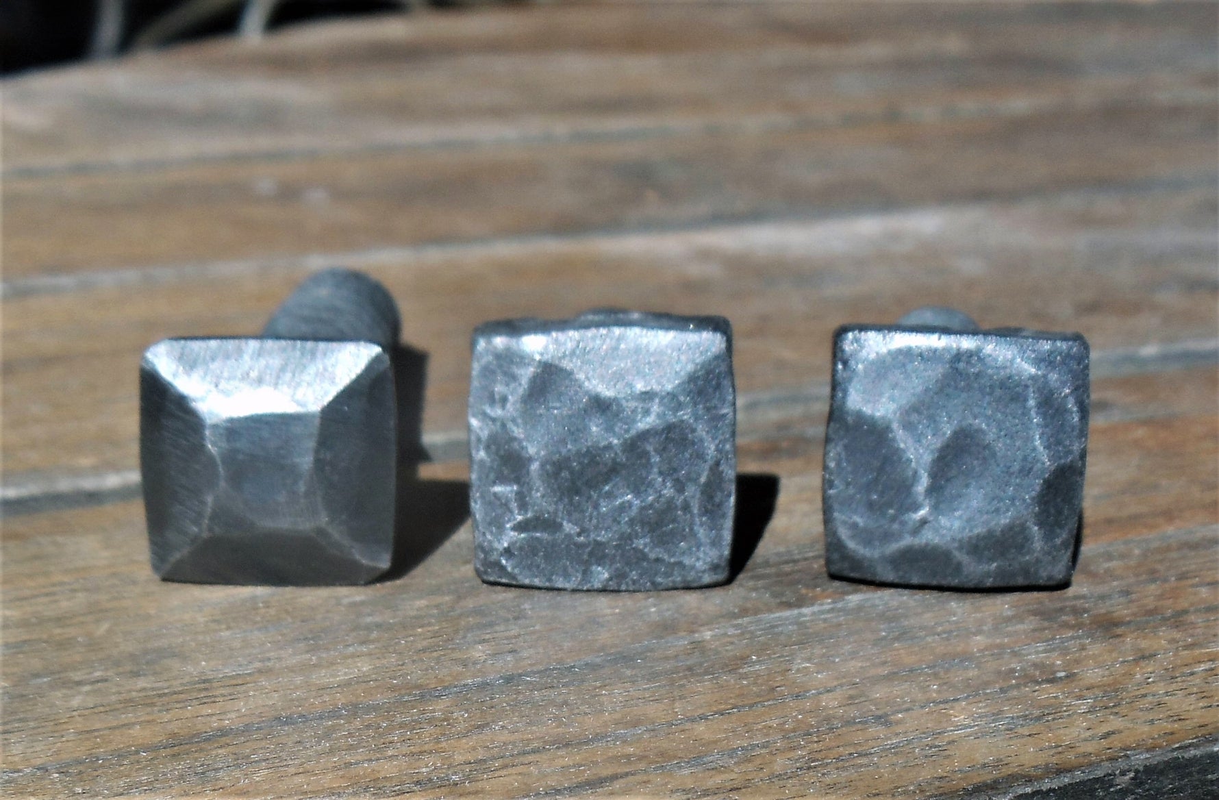 Three examples of hand hammered square head bolts Pyramid Hammered, disstressed, and hammered with a raw finish on a wood background.