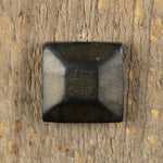 A pyramid hammered square head bolt that is painted black and finished with an aged gold patina installed on a wood background.
