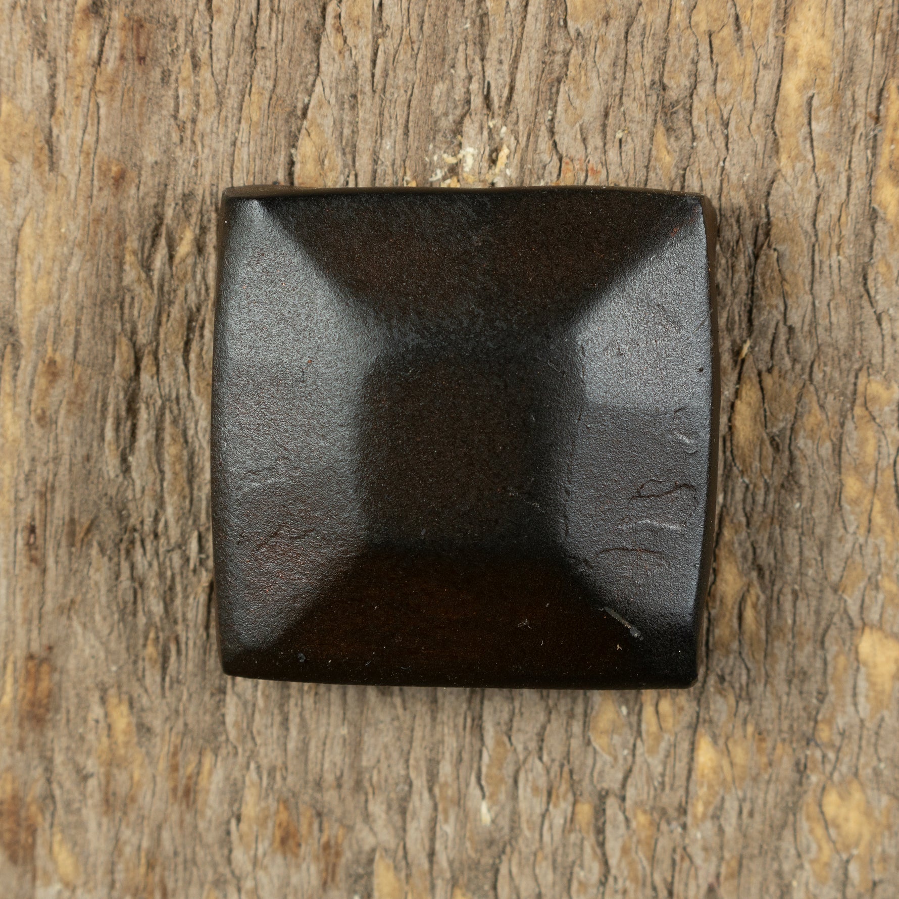 A pyramid hammered square head bolt that is painted black with a rusty metal patina installed on a wood background.