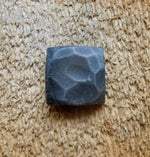 A top view of a hand hammered square head lag that has been painted black with an aged pewter patina finish screwed into a piece of wood as the background.